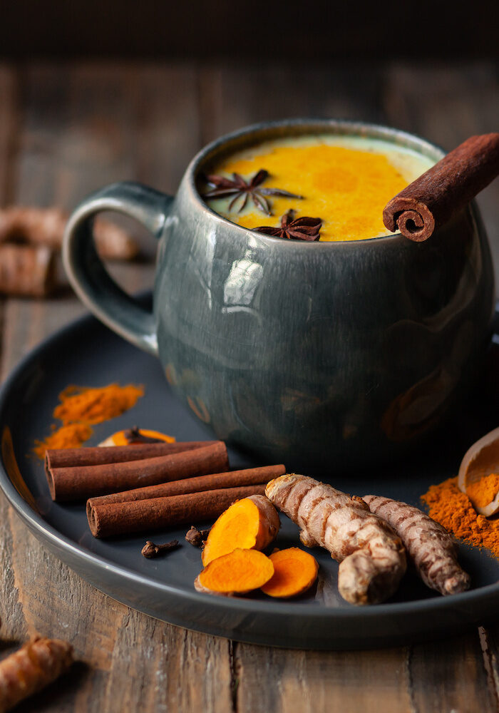 Golden milk in grey mug. Turmeric latte made with curcuma, cinnamon, anise. Healthy hot winter drink, natural, organic beverage. Close up, front view. Wooden rustic background. Raw roots as decor